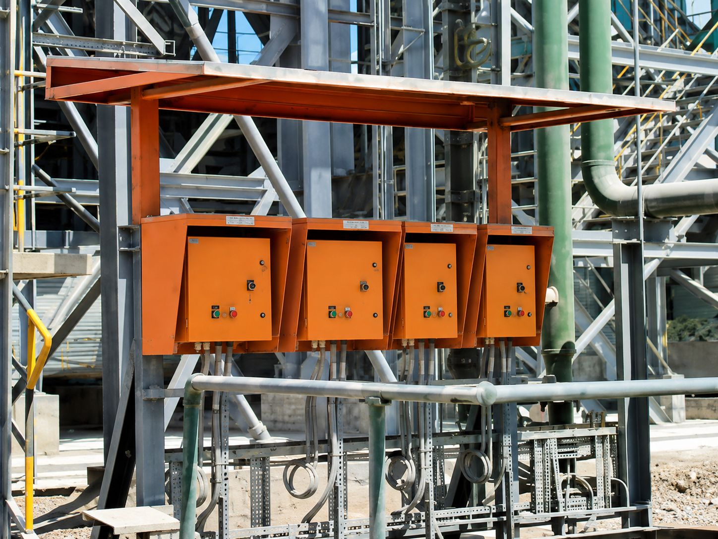 Four orange control panels under a steel weather canopy at a cement plant, with conduit dropping to equipment below